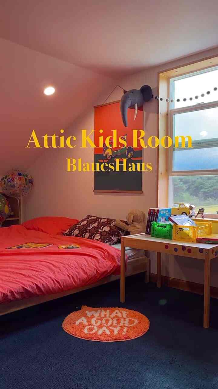 Orange-toned poster and red bed in attic kids' room featuring a wooden desk by a bright window, creating a cozy natural-lit space