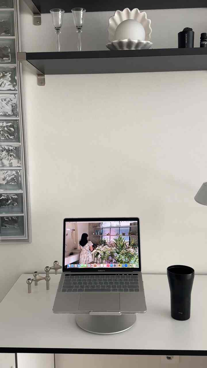 シルバーとホワイトの色調のホームオフィス、中央にMacBook Proとノートパソコンスタンドが配置され、隣に黒色のタンブラーが置かれた整ったデスク